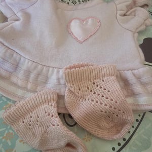 May include: A light pink doll outfit, including a top with a heart applique and ruffled hem, and a pair of matching socks. The top has short, puffed sleeves. The socks have a decorative openwork design.