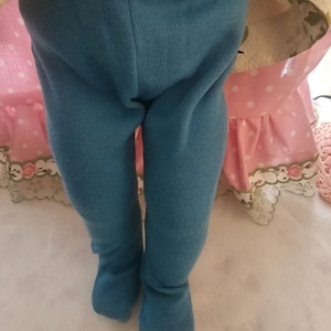 May include: Teal-colored doll leggings with attached feet. The leggings are made of a ribbed knit fabric. The doll is wearing a pink polka-dot dress with a floral pattern on the bodice.