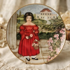 May include: A round decorative ceramic plate with a gold rim featuring a painted scene of a child in a red dress holding a rose and a basket of flowers. The background shows a large stone house, green landscaping, and pink rose bushes.