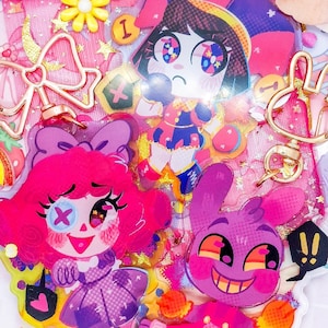 Puede incluir: Un conjunto de cuatro amuletos de personajes coloridos con acentos rosa, morado y rojo. Los amuletos están unidos a llaveros dorados con una bolsa de malla rosa. Los amuletos representan a un bufón, una niña con un lazo rosa, una niña con un vestido rosa y un monstruo morado.