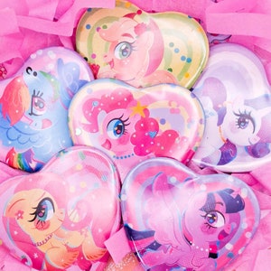 Op de afbeelding: Zes hartvormige pins met My Little Pony-personages. Elke pin heeft een ander personage met een kleurrijke achtergrond. De pins zijn in een cirkel op een roze achtergrond gerangschikt.