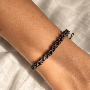 Könnte beinhalten: Ein dunkelgraues Makramee-Armband mit einem sich wiederholenden Knotenmuster. Das Armband besteht aus einer dünnen Kordel und verfügt über verstellbare Zugbänder an den Enden, die mit kleinen Metallperlen für einen sicheren Sitz versehen sind.