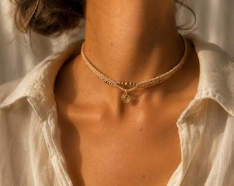 Può includere: Una collana girocollo in corda beige dal design a doppio strato con piccole perline in metallo dorato e un ciondolo circolare centrale in oro con dettagli a cerchi concentrici. La collana è indossata al collo sopra una camicia bianca.
