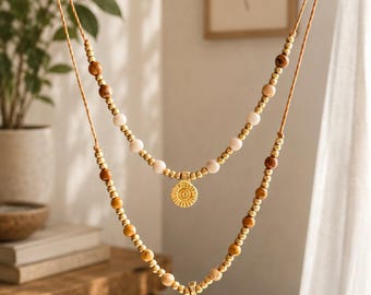 Può includere: Una collana a due fili composta da perline marroni, color cuoio e oro metallizzato. Il filo più corto include perline chiare e un piccolo ciondolo circolare dorato, mentre il filo più lungo presenta un ciondolo dorato a forma di sole.