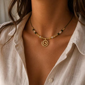 May include: A necklace featuring a gold-toned spiral pendant suspended from a cord. The cord is adorned with a series of small, alternating beads in shades of translucent white, yellow, and dark green, interspersed with tiny metallic gold-toned spacers.