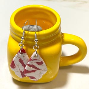 Könnte beinhalten: Handgefertigte Ohrringe mit einem einzigartigen Design. Die Ohrringe haben eine weiße Basis mit roten und goldenen Akzenten. Sie werden in einer leuchtend gelben Tasse präsentiert. Die Ohrringe haben silberfarbene Haken.