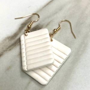 May include: A pair of white, rectangular earrings with a gold hook. The earrings have a textured, ribbed surface.