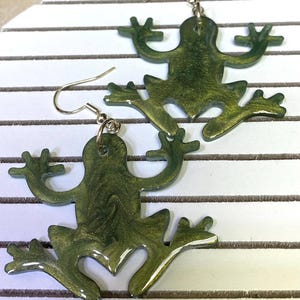 May include: A pair of green frog-shaped earrings with a gold shimmer finish. The earrings are made of resin and have a hook fastening.
