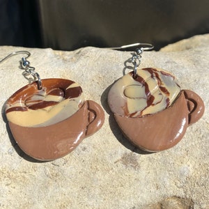 Puede incluir: Un par de pendientes de taza de café de color marrón y beige con un patrón de remolino. Los pendientes están hechos de arcilla polimérica y tienen ganchos de plata.