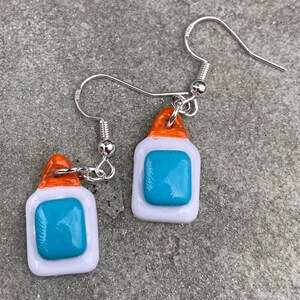 May include: Handmade earrings with a miniature glue bottle design. Each earring has a white body, an orange cap, and a turquoise blue square. The earrings are suspended from silver-coloured fish hook ear wires.