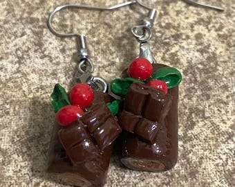 May include: A pair of handmade earrings featuring miniature chocolate log designs. Each earring has a dark brown cylindrical base, topped with red cherry accents, green leaves, and chocolate bar details. The earrings hang from silver-colored hooks.