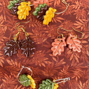 May include: Three pairs of earrings featuring autumn leaves in various shades of brown, orange, and green. The leaves are made of polymer clay and are attached to gold and silver hoops and hooks.