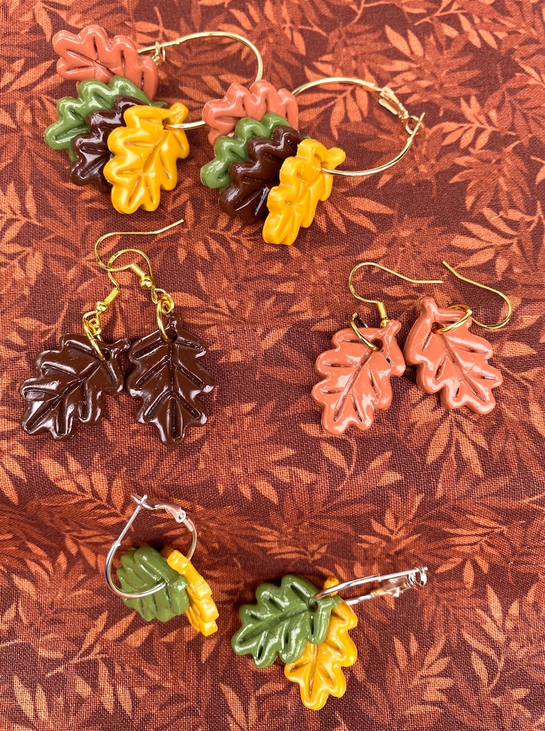 May include: Three pairs of earrings featuring autumn leaves in various shades of brown, orange, and green. The leaves are made of polymer clay and are attached to gold and silver hoops and hooks.