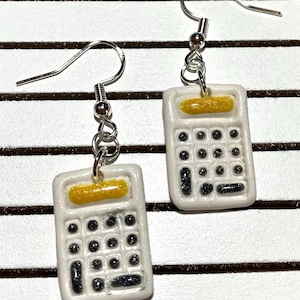 Puede incluir: Un par de pendientes de calculadora blancos con una pantalla amarilla y botones negros. Los pendientes están hechos de arcilla polimérica y tienen ganchos plateados.