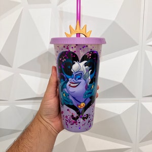 May include: Purple tumbler with a lid, straw, and a gold crown-shaped topper. The tumbler features a design of Ursula from The Little Mermaid, with a purple and black color scheme and glitter accents.