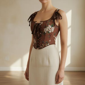 May include: A brown corset top with a floral pattern and brown ribbon ties. The top has brown shoulder straps with bows. The corset is paired with a cream-colored skirt.