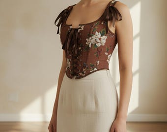 May include: A brown corset top with a floral pattern and brown ribbon ties. The top has brown shoulder straps with bows. The corset is paired with a cream-colored skirt.