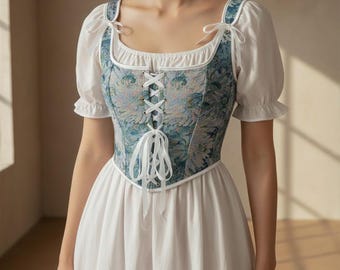 Könnte beinhalten: Weißes Kleid mit quadratischem Ausschnitt und kurzen, gepufften Ärmeln. Das Oberteil hat ein blau-grünes Blumenmuster mit weißer Schnürung vorne. Das Kleid hat eine Vintage- und romantische Ästhetik.