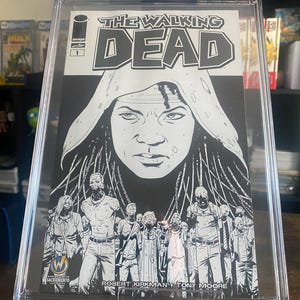 May include: A black and white comic book cover for The Walking Dead #1. The cover features a woman with long, dark hair and a hooded jacket. The title "The Walking Dead" is written in large, bold letters at the top of the cover. The cover also includes the text "Image Comics, 6/15" and "Robert Kirkman Tony Moore".