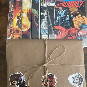 Pu&ograve; includere: Una pila di sei fumetti in buste protettive di plastica, inclusi titoli come What If The Punisher Had Killed Daredevil, dietro un pacchetto di carta marrone legato con spago e decorato con tre adesivi di personaggi.