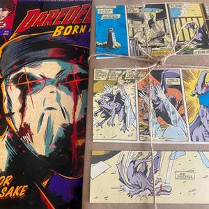 Pu&ograve; includere: Un fumetto Daredevil Born Again con un'illustrazione scura di una figura mascherata con occhi rossi, posizionato accanto a un pacchetto rettangolare avvolto in carta marrone e legato con spago, decorato con diversi pannelli di fumetti.
