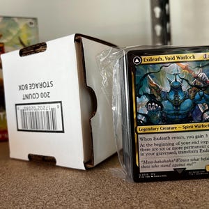 May include: A pack of trading cards with a blue and black fantasy character, labelled "Exdeath, Void Warlock." A white 200-count storage box is visible, alongside a box of Magic: The Gathering cards.
