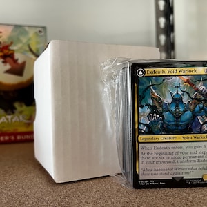 May include: A stack of Magic: The Gathering cards, featuring the "Exdeath, Void Warlock" card. The card has a blue and gold colour scheme with detailed artwork of a fantasy creature. The card is in a plastic sleeve, with a white box and a game box in the background.