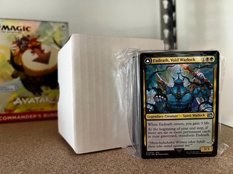 May include: A stack of Magic: The Gathering cards, featuring the "Exdeath, Void Warlock" card. The card has a blue and gold colour scheme with detailed artwork of a fantasy creature. The card is in a plastic sleeve, with a white box and a game box in the background.