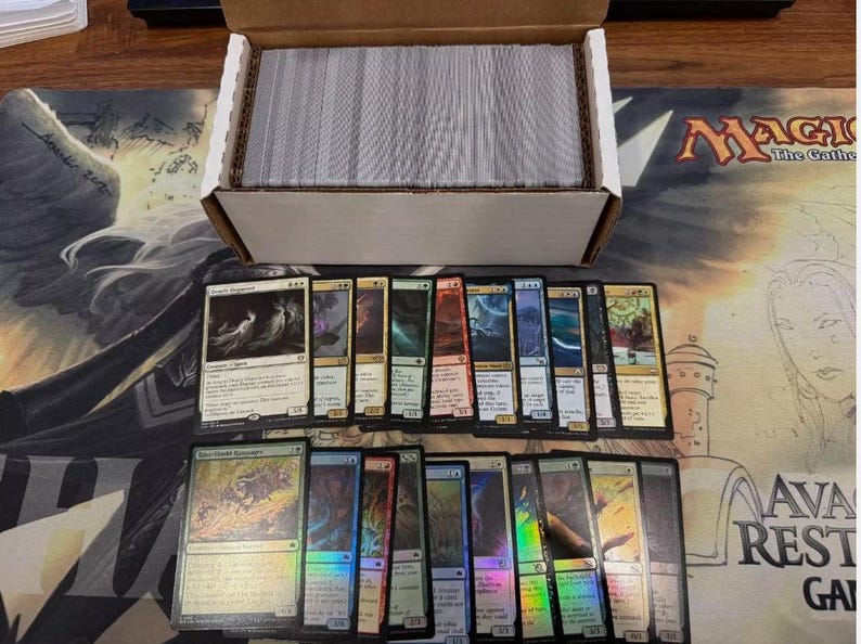 May include: A collection of Magic: The Gathering trading cards, some with holographic foil, arranged on a playmat. A cardboard box filled with more cards sits above the display. The cards feature various colors and artwork.