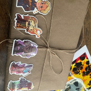 Pu&ograve; includere: Un pacchetto rettangolare in carta marrone legato con spago di juta sottile, decorato con cinque adesivi colorati di personaggi dei cartoni animati, accanto a una stampa rettangolare separata che mostra un teschio e immagini di fuoco.