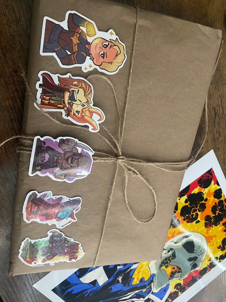 Pu&ograve; includere: Un pacchetto rettangolare in carta marrone legato con spago di juta sottile, decorato con cinque adesivi colorati di personaggi dei cartoni animati, accanto a una stampa rettangolare separata che mostra un teschio e immagini di fuoco.
