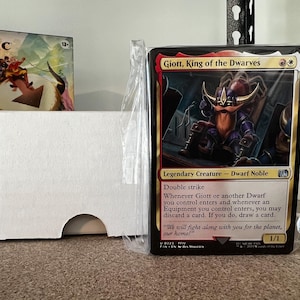 May include: A Magic: The Gathering card featuring Giott, King of the Dwarves, a Dwarf Noble with double strike. The card is in a protective sleeve, next to a white card box and a game box.