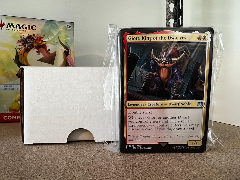 May include: A Magic: The Gathering card featuring Giott, King of the Dwarves, a Dwarf Noble with double strike. The card is in a protective sleeve, next to a white card box and a game box.