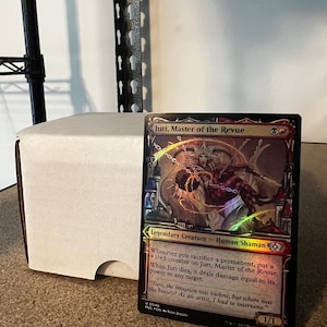 May include: A holographic trading card featuring "Juri, Master of the Revue," a Legendary Creature Human Shaman. The card has a colourful design with text describing its abilities and a quote. A white card storage box is in the background.