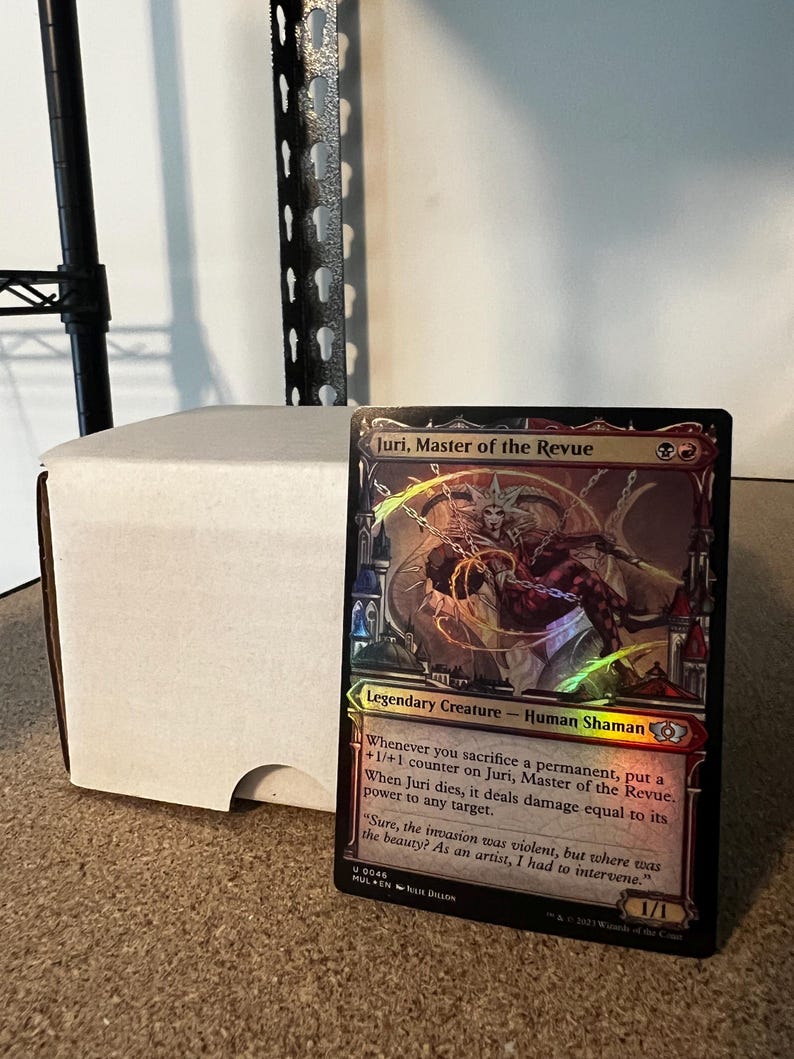 May include: A holographic trading card featuring "Juri, Master of the Revue," a Legendary Creature Human Shaman. The card has a colourful design with text describing its abilities and a quote. A white card storage box is in the background.