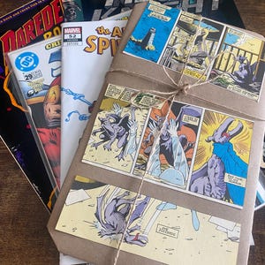 Pu&ograve; includere: Una pila di fumetti vintage tra cui Moon Knight, Daredevil e The Amazing Spider-Man, sormontata da un pacchetto avvolto in carta marrone legato con spago e decorato con pannelli a fumetti colorati raffiguranti creature simili a gargoyle.