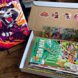 May include: A comic book box filled with comic books, trading cards, and stickers. The box contains a stack of comic books, including an "Iron Man" comic. A Rocket Raccoon art print is displayed on a stand. Stickers of Marvel characters are visible.