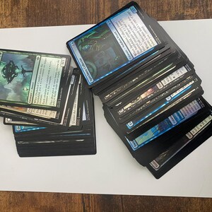 May include: Stacks of Magic: The Gathering trading cards, featuring various card designs and colors. The cards are arranged in two separate piles on a white surface. Some cards have text and illustrations.