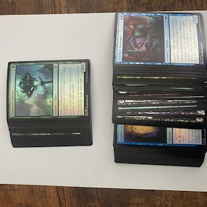 May include: Two stacks of Magic: The Gathering trading cards. One stack features a green card with the text "Duskshell Crawler." The other stack has blue cards, including "Stitched Assistant." Cards are on a white surface.