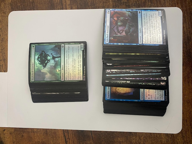 May include: Two stacks of Magic: The Gathering trading cards. One stack features a green card with the text "Duskshell Crawler." The other stack has blue cards, including "Stitched Assistant." Cards are on a white surface.