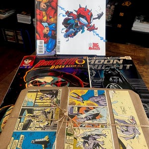 Pu&ograve; includere: Una collezione di fumetti tra cui The Amazing Spider-Man, Daredevil Born Again e Moon Knight, insieme a un pacchetto avvolto in carta marrone legato con spago, con vignette di fumetti stampate sulla superficie anteriore.