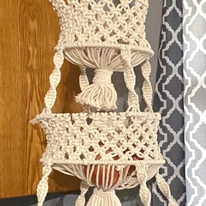 May include: A three-tiered macrame hanging planter with a white rope and a wooden ring at the top. The planter has three baskets, each with a different pattern. The baskets are empty.