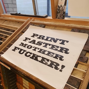 May include: A white sheet of paper with the words "PRINT FASTER MOTHER FUCKER!" printed in bold, black letters. The paper rests on a wooden printing press with various letter blocks.