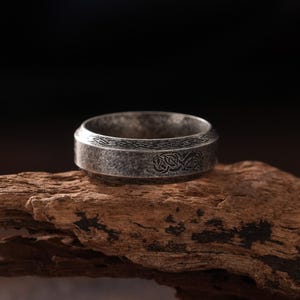 May include: A silver-toned metal ring with a distressed, weathered finish and a dark, engraved Celtic knot pattern encircling the band. The ring features a flat, wide profile with beveled edges, showcasing intricate, repeating knotwork details.