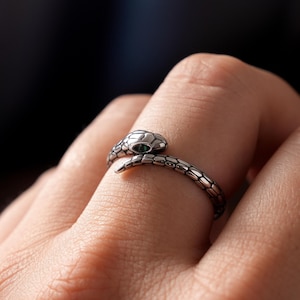 May include: A silver-toned metal ring shaped like a coiled snake with textured scales wrapping around the finger. The snake head features a small, dark green stone set as an eye, with the body forming an open-ended band design.