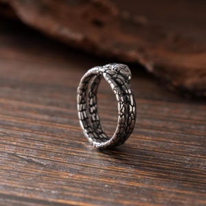 May include: A silver-toned metal ring shaped like a coiled snake. The band features a detailed, textured scale pattern covering the entire surface, with the snake's head resting on the top of the band, completing the circular form of the ring.