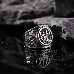 Può includere: Un anello d'argento con un design a forma di impronta di zampa d'orso. L'anello è dettagliato con un motivo a nodo celtico e ha una finitura testurizzata.
