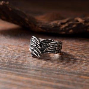 Può includere: Un anello in metallo color argento con un design dettagliato di piume sovrapposte che avvolgono l'anello. La superficie presenta una finitura scura e ossidata nelle aree incavate per mettere in risalto la trama e le linee delle piume.