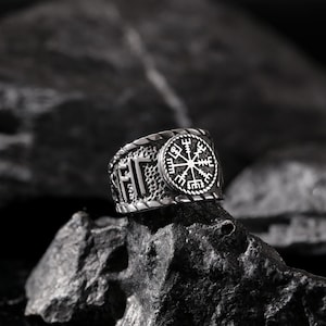 May include: A silver ring with a textured band and a circular design featuring a compass with eight points. The compass is surrounded by a circle with a pattern of dots and lines.