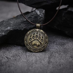 May include: A round, antique brass-toned pendant featuring a relief design of a bear silhouette inside a large paw print. The pendant has a textured, decorative border and is suspended from a thin, dark brown cord necklace.